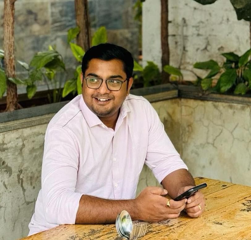 Ajitesh Akula smiling, wearing a light pink shirt and glasses, seated at a wooden table with greenery in the background — Knoxville Telugu Association leadership